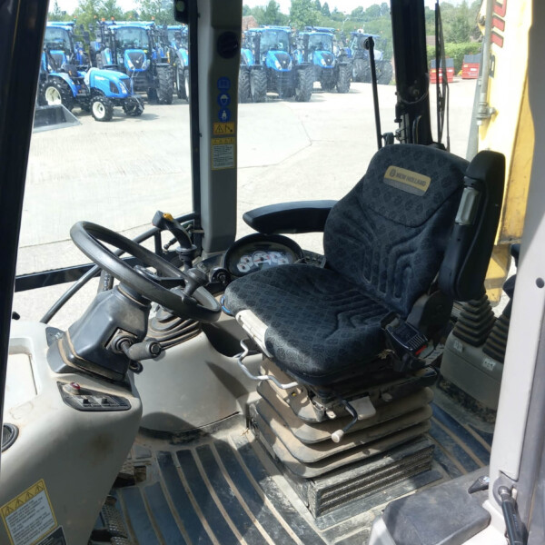 Inside tractor cab showing steering wheel and driver's seat, with multiple blue tractors parked outside in a lot