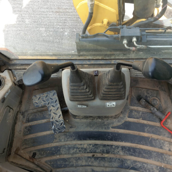 Cab view of a construction excavator with two joysticks, pedals, and dusty interior.