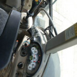 Operator's cab of a heavy construction machine showing the gear lever, joystick, and dashboard with multiple gauges and switches. Through the window you can see the outdoor ground and a warning label nearby.