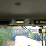 Truck cab interior with overhead light, vents, and a Pioneer touchscreen stereo; outside a street with blue barriers and greenery.