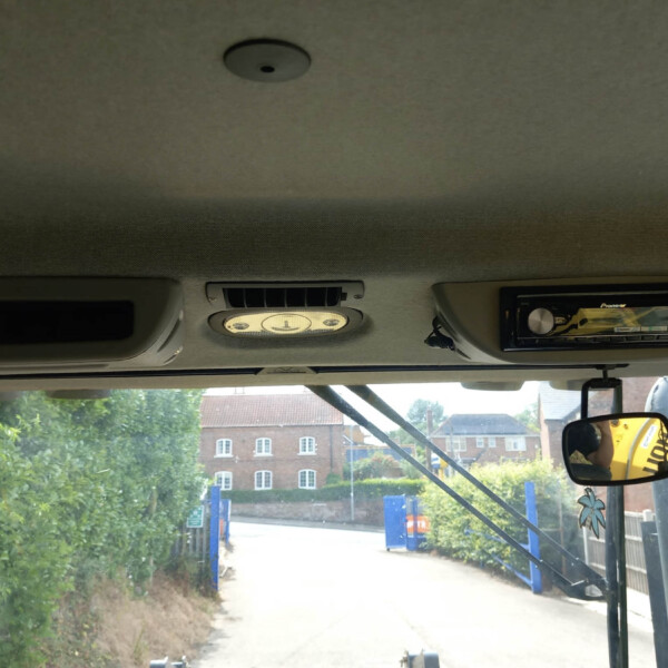 Truck cab interior with overhead light, vents, and a Pioneer touchscreen stereo; outside a street with blue barriers and greenery.