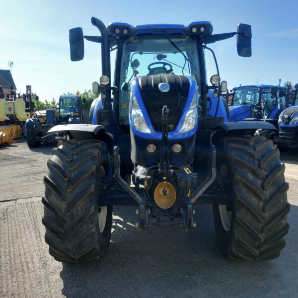 Front view of a blue tractor in a yard with rows of blue tractors and yellow equipment nearby under a clear sky.