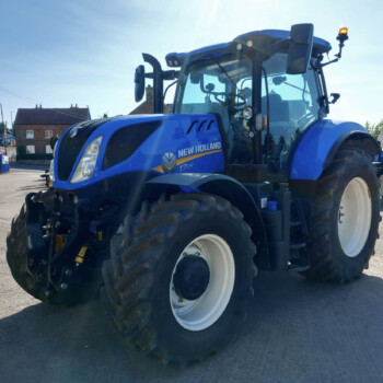 Blue New Holland tractor parked on a street, with houses in the background and clear sunny sky.