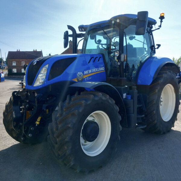 Blue New Holland tractor parked on a street, with houses in the background and clear sunny sky.