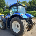 Blue New Holland tractor front with oversized rear wheel, parked in a lot near a dealership sign that reads 'New Holland' and 'Russells.'