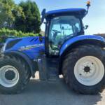 Blue New Holland tractor viewed from the side, showing large front and rear tires, cabin, and a sunny outdoor lot.