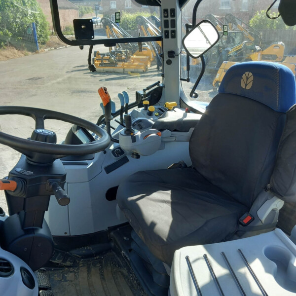 Interior of a heavy machinery cab (tractor) showing steering wheel, multiple levers, pedals and control panel, with construction equipment visible through the windshield outside
