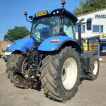 Front view of a blue New Holland tractor with oversized tires parked outside a farm equipment dealership, orange warning lights on top. California? (note: keep concise)