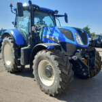 Blue New Holland tractors lined up in a parking lot on a sunny day, close-up of a front tractor with large tires in the foreground.