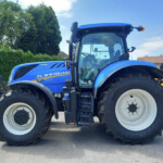 Blue New Holland T7.225 tractor, side view with large rear tires, parked on a paved lot with hedges and buildings in the background