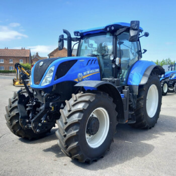 Blue New Holland tractor in foreground with other blue tractors lined up behind in a rural lot; houses in background under a clear sky