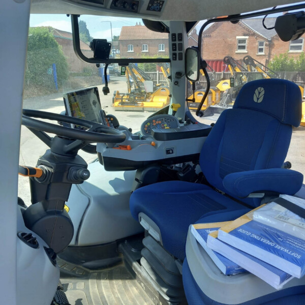 Driver's cab of a blue tractor with a steering wheel, dashboard controls, and a blue seat; view of yard machinery outside the window