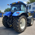 Large blue tractor on a dealership lot with other blue machinery and a New Holland sign in the background.