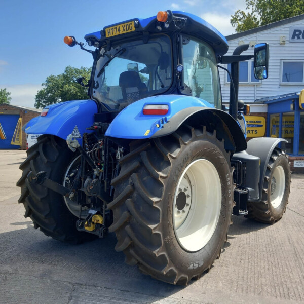 Large blue tractor on a dealership lot with other blue machinery and a New Holland sign in the background.