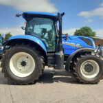 Blue New Holland tractor with oversized tires parked outdoors, service sign in background
