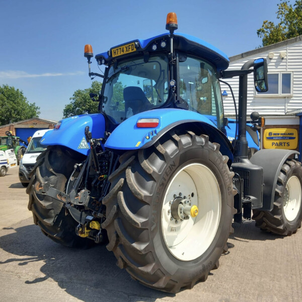 Blue agricultural tractor with oversized rear tires, orange beacon lights, parked in a lot beside a dealership building with other vehicles in the background