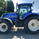 Blue New Holland T7.260 tractor parked outdoors, close-up of the front wheel and cab with an orange beacon on top, trees and buildings in the background
