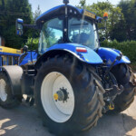 Front-right view of a blue New Holland tractor with oversized white wheels and a cab, parked outdoors near a dealership sign.