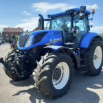 Blue New Holland T7.260 tractor parked in a yard with other tractors and red-brick houses in the background on a sunny day.
