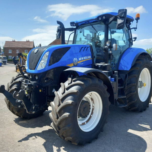 Blue New Holland T7.260 tractor parked in a yard with other tractors and red-brick houses in the background on a sunny day.