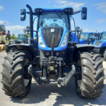 Front view of a blue agricultural tractor with oversized tires, parked among other blue tractors and equipment outdoors under a bright sky.