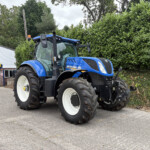 Blue New Holland tractor parked outside a dealership, with a green hedge and a white building in the background.