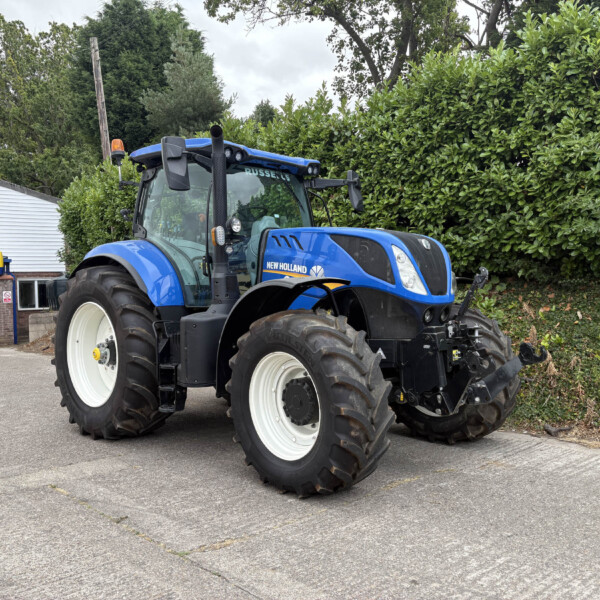 Blue New Holland tractor parked outside a dealership, with a green hedge and a white building in the background.