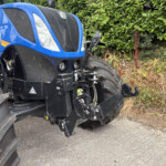 Close-up of a blue New Holland tractor facing left beside a green hedge on a paved surface.