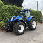 Blue New Holland tractor parked on a street beside a tall green hedge under a cloudy sky.