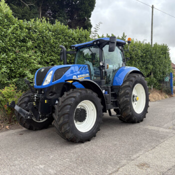 Blue New Holland tractor parked on a street beside a tall green hedge under a cloudy sky.