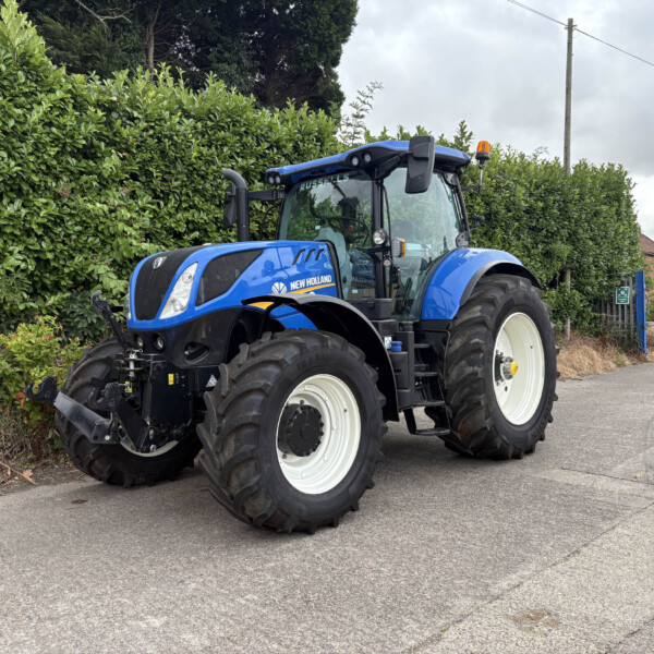 Blue New Holland tractor parked on a street beside a tall green hedge under a cloudy sky.
