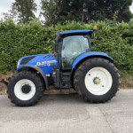 Blue New Holland T7-260 tractor parked beside a tall green hedge, orange beacon on the roof, front wheels visible.