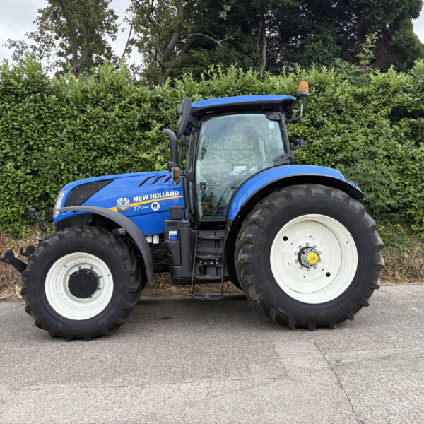 Blue New Holland T7-260 tractor parked beside a tall green hedge, orange beacon on the roof, front wheels visible.