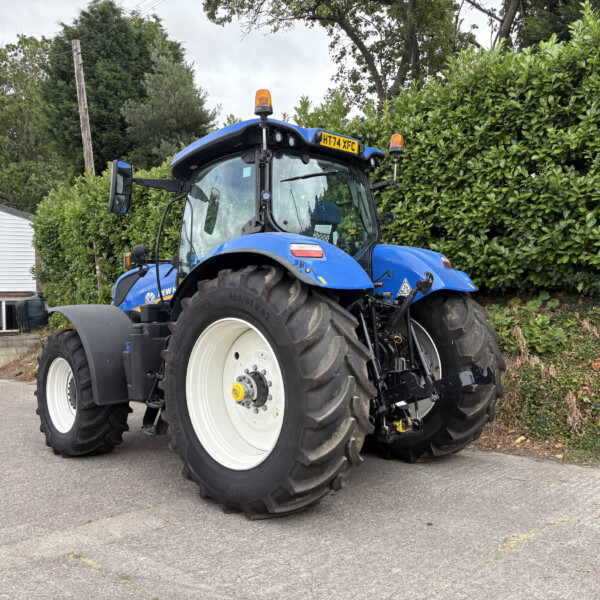 Blue agricultural tractor with large white wheels parked in a lot beside a hedge and a building with a yellow 'SELLS' sign in the background.