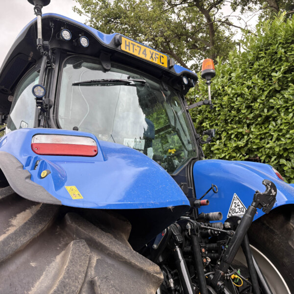 Blue tractor cab close-up with oversized front tires and orange beacon on the roof; hedge in background and license plate HT74 XFC visible.