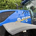 Close-up of a blue New Holland tractor hood and cab with the model name T7.260 and logo, hedge in the background.