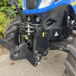 Close-up of the front left side of a blue tractor showing the hitch, hydraulic linkage, and large rugged tires.