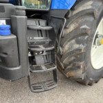 Close-up of a tractor's step area with black plastic steps, blue body panels, and a large muddy rear tire with a yellow hub.