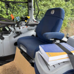Interior of a tractor cab: blue driver's seat, control levers, and a cardboard box on the floor with a boxed manual on the seat still strapped