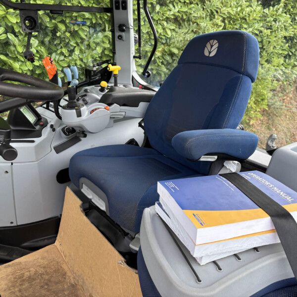 Interior of a tractor cab: blue driver's seat, control levers, and a cardboard box on the floor with a boxed manual on the seat still strapped
