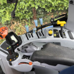 Interior of a tractor cabin showing multiple levers, a yellow control knob, and an orange handle, with green foliage outside the window.