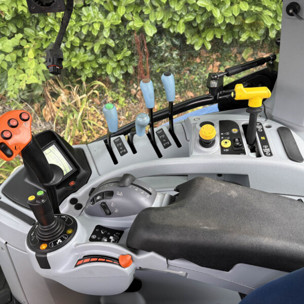 Interior of a tractor cabin showing multiple levers, a yellow control knob, and an orange handle, with green foliage outside the window.