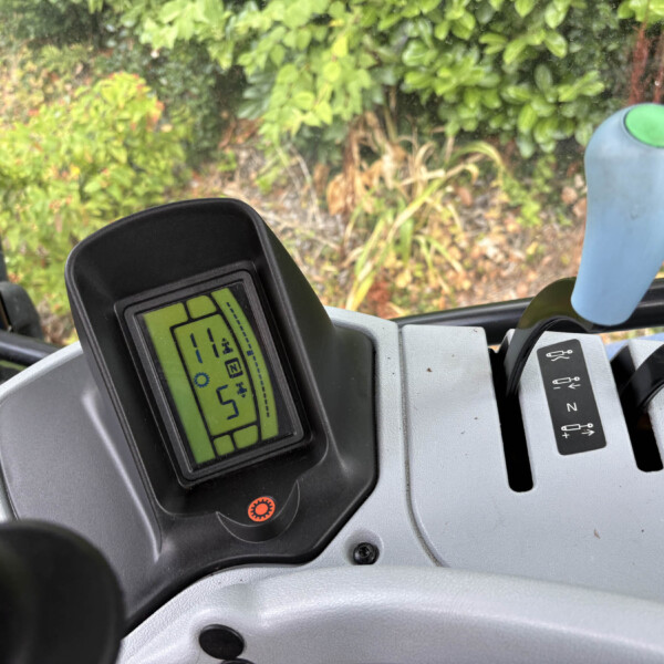 Close-up of a tractor cabin dashboard with a green digital display, a blue joystick with a green top, and garden foliage outside the window.