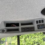 Interior ceiling of a vehicle showing an overhead control panel with buttons, an empty open compartment, a circular speaker, and window views of greenery outside.