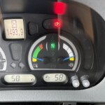 Close-up of a car dashboard: left dial shows speedometer, center digital readout reads 1493.9 with a small icon above, and a red parking brake indicator is illuminated; other gauges and colored indicators are visible around the bezel.