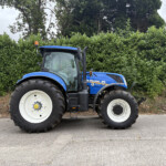 Blue New Holland tractor parked on a road beside a tall green hedge and trees, with an orange beacon on top of the cab.