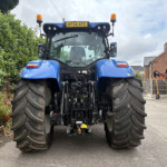 Rear view of a large blue tractor with wide tires, orange beacon lights, and equipment in a yard along a hedge.