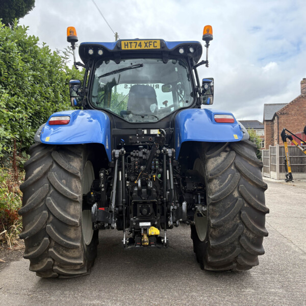 Rear view of a large blue tractor with wide tires, orange beacon lights, and equipment in a yard along a hedge.