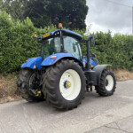 Blue agricultural tractor with oversized rear tires parked beside a tall hedge on a paved road; orange beacon lights on top.