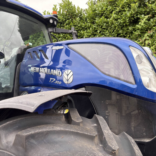 Blue New Holland T7.300 tractor front with large headlights and chrome grille, hedge in background.